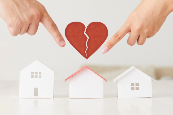 Hands point at a broken red heart above three small white paper houses, symbolizing relationship breakdown and division. The mood is tense and somber.