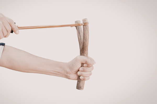 A hand holding a wooden slingshot, pulling back the elastic band, poised to shoot. The background is plain, emphasizing focus and tension.