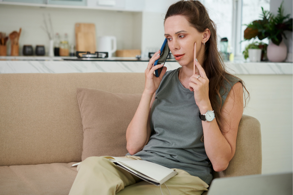 A woman on a beige sofa in a modern kitchen talks on her phone, holding a pen and an open notebook. She appears focused and contemplative.