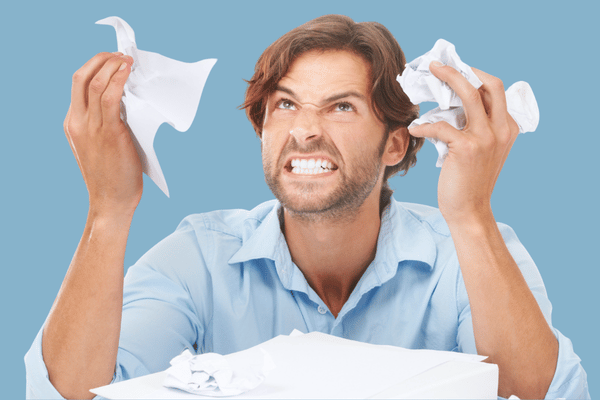 Frustrated man in a light blue shirt grimaces while clutching crumpled tissues, surrounded by paper, suggesting stress, anger, or a messy cleanup.